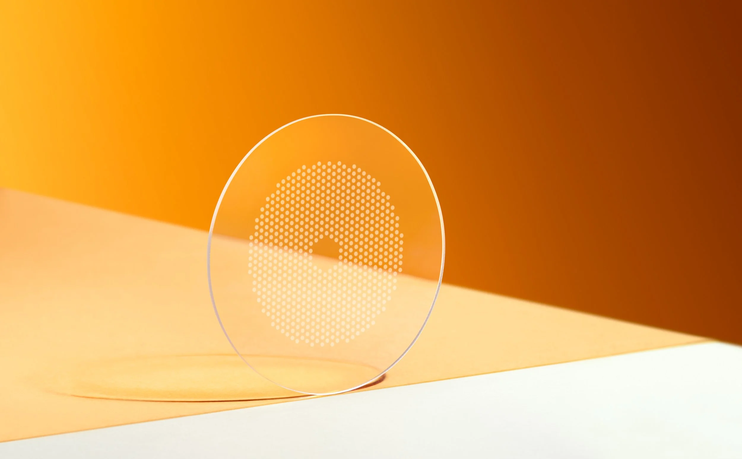 miyo-smart-contact-lens-orange-product-shot. Transparent circular smart contact lens with a white dotted pattern, standing upright on a softly lit orange surface against a warm orange gradient background. |

Hoya Miyo Smart
