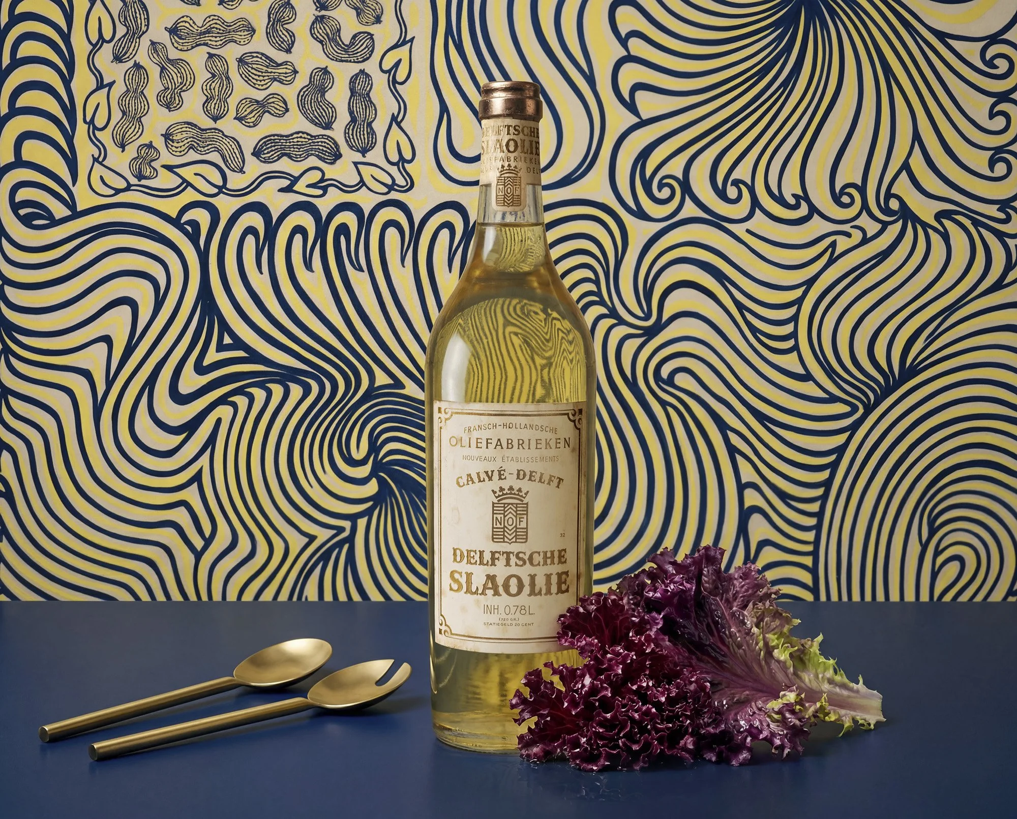 Vintage Delftsche Slaolie bottle with red leaf lettuce and gold salad servers on a blue table, set against a blue and yellow illustrated background.