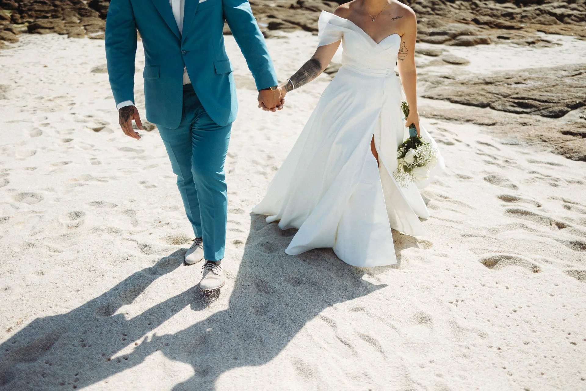 Ein Paar in Hochzeitskleidung hält Händchen an einem Strand, die Frau trägt ein weißes Brautkleid und hält einen Blumenstrauß, der Mann trägt einen blauen Anzug.