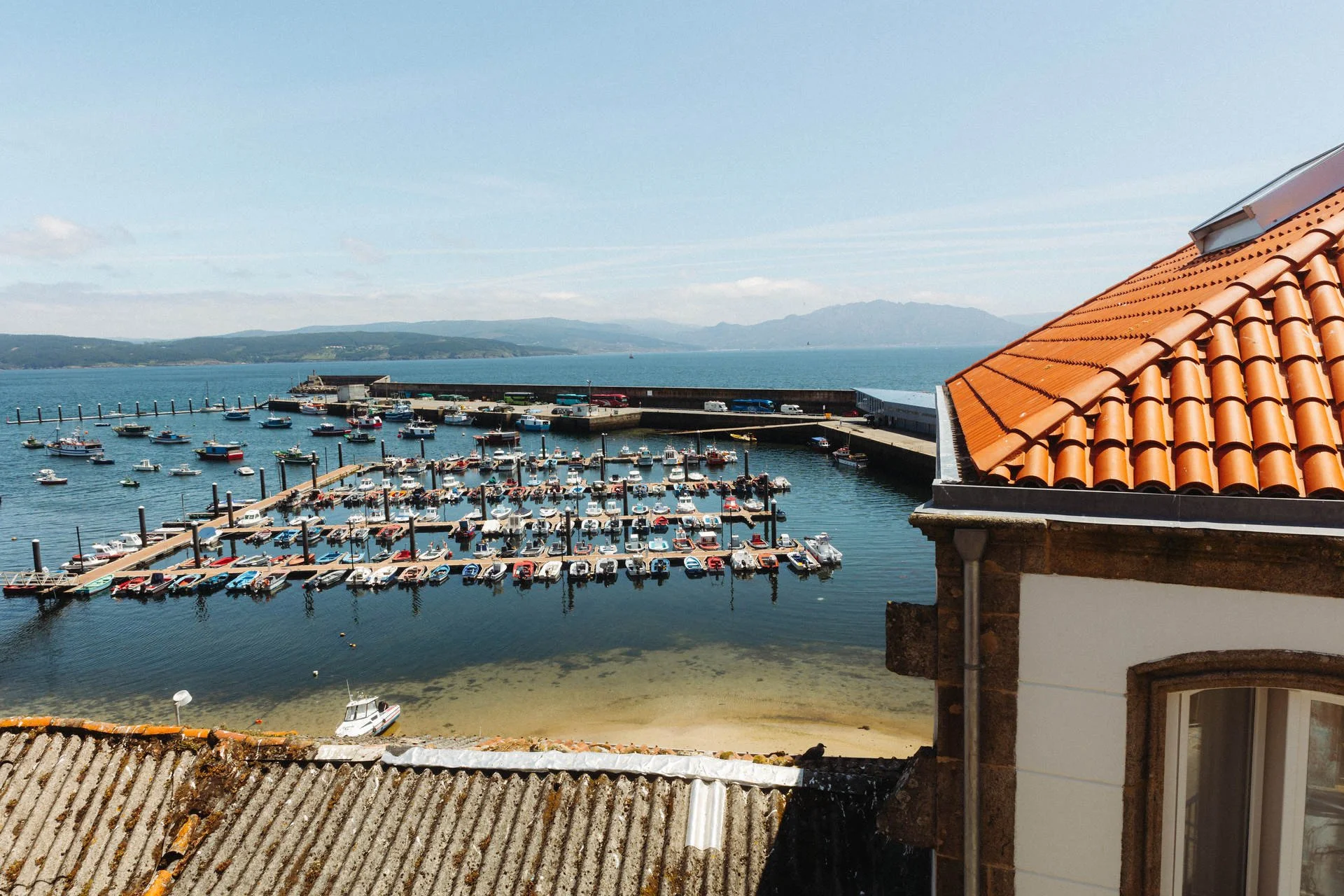 Blick auf einen Hafen mit vielen Booten, umgeben von Wasser und Bergen im Hintergrund, mit roten Ziegeldächern im Vordergrund.