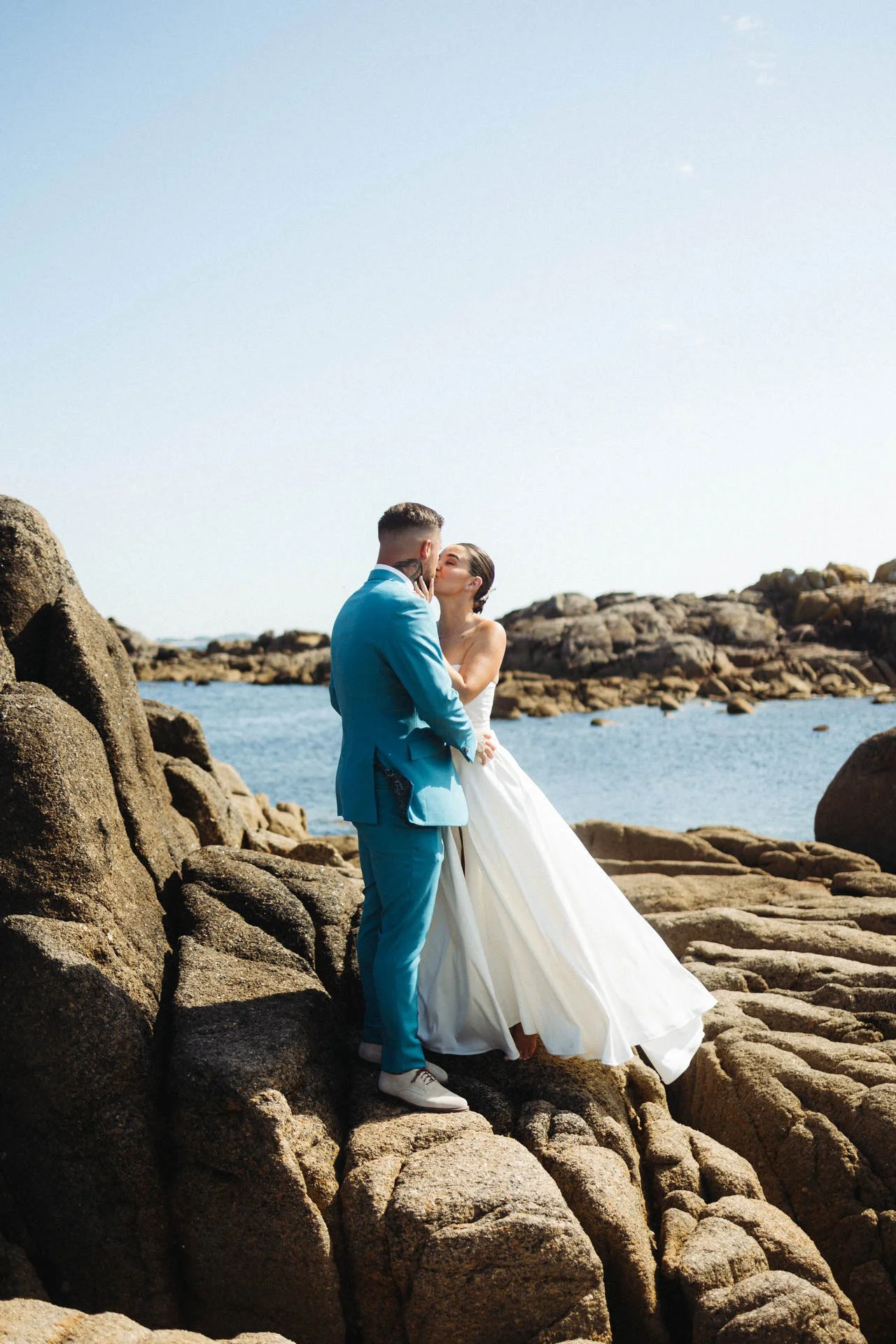 Ein Brautpaar küsst sich an Felsen am Meer bei sonnigem Himmel.