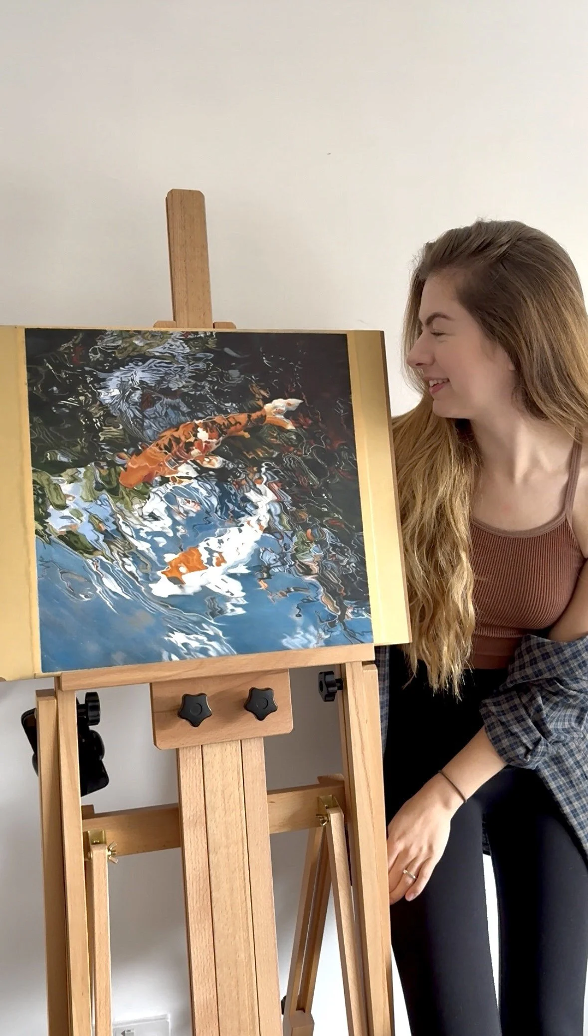 A young woman with long blonde hair looking at a colorful, impressionistic drawing of koi fish in water on a wooden easel.