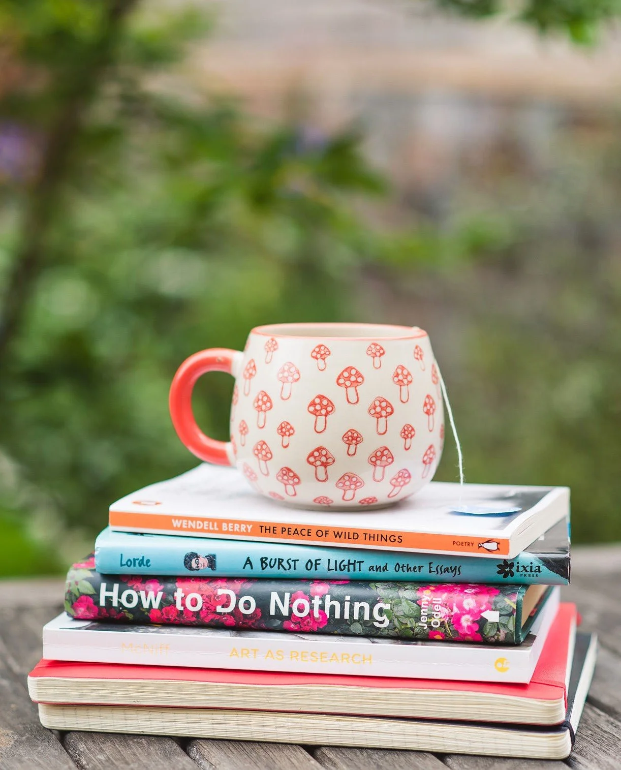 A stack of books with a red mug on top, which has a playful red mushroom pattern. The books include 'The Peace of Wild Things' by Wendell Berry, 'Art As Research' and 'How to do Nothing' by Jenny Odell