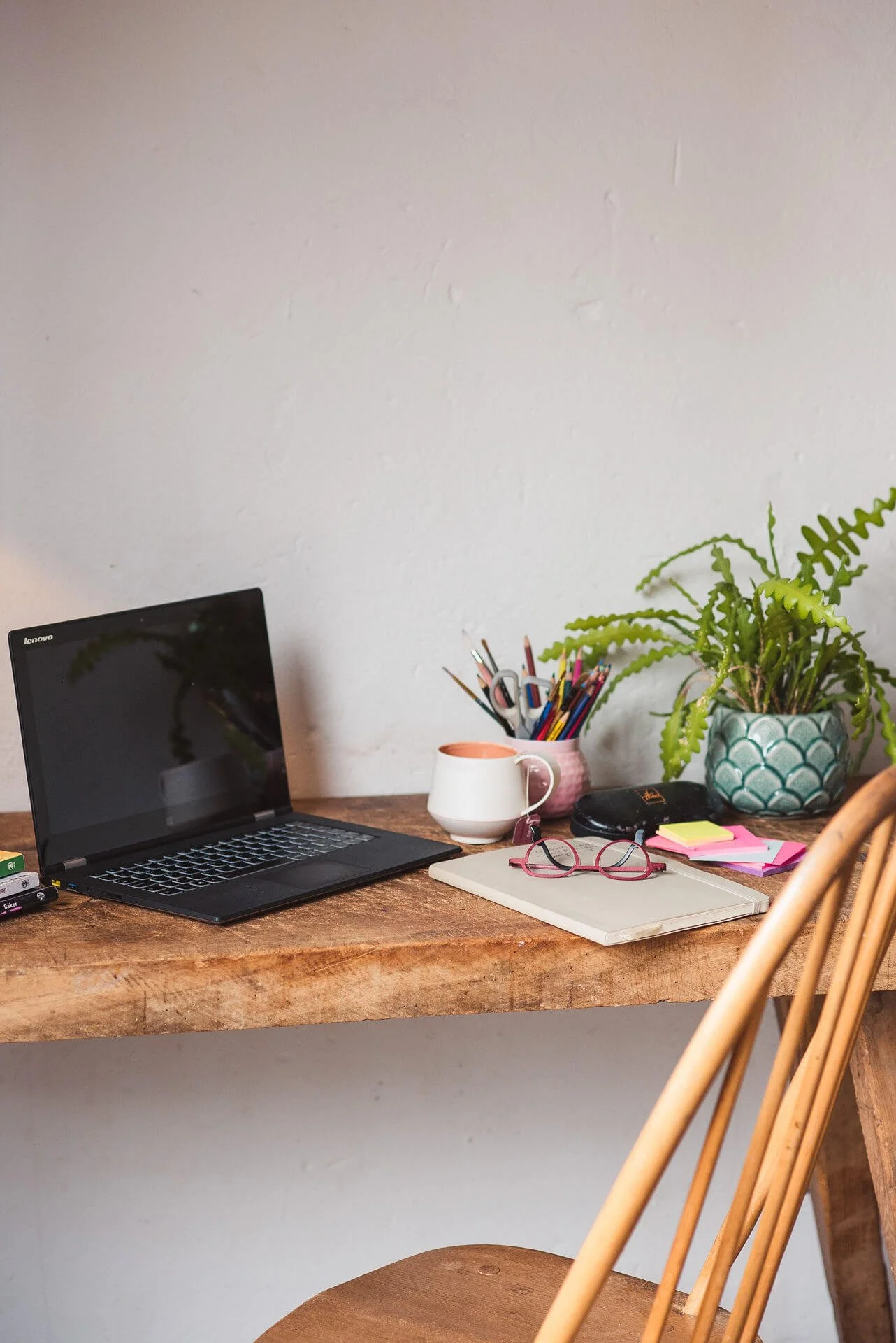 A cosy workspace, with a wooden desk and chair, laptop, notebook, pen pot, mug & plant