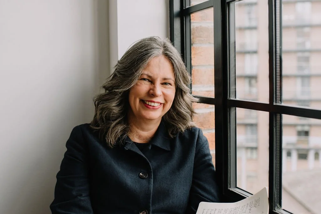 Jane Willis sits infront of a window holding a journal, smiling directly at the camera
