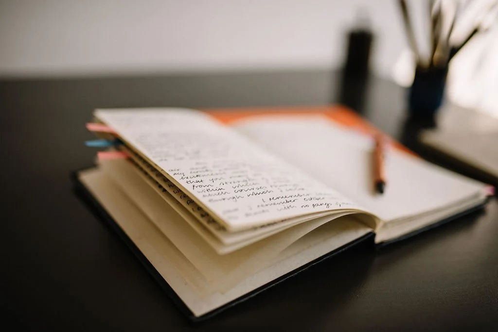 Open notebook with handwritten notes, a pen on top, on a dark desk with some objects in the background.