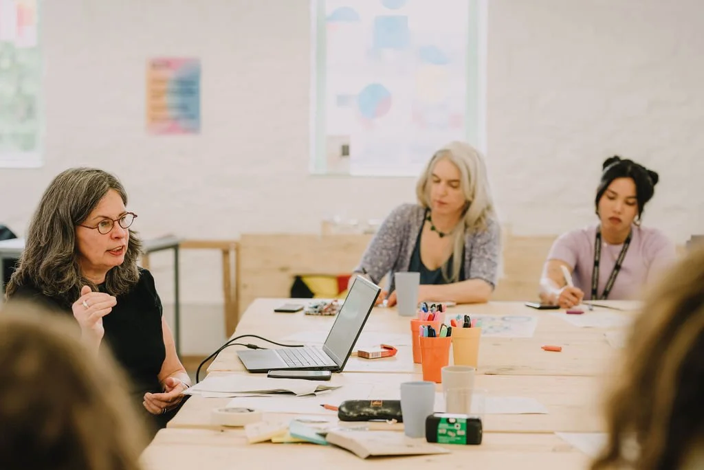 Jane Willis sits at a table leading a group workshop for arts & health professionals