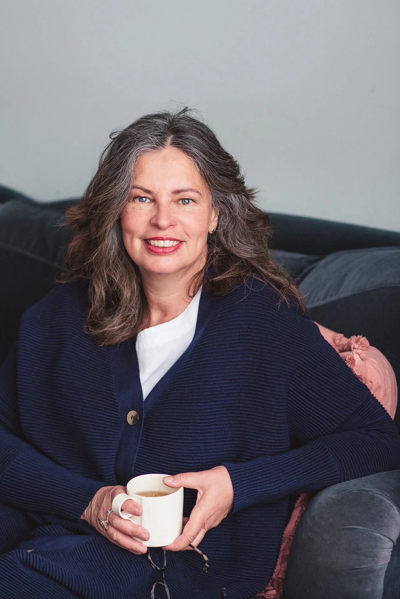 A headshot of Jane Willis, coach, mentor and trainer. Jaane sits looking at the camera holding a mug of tea, a white woman with dark hair, wearing a blue sweater.
