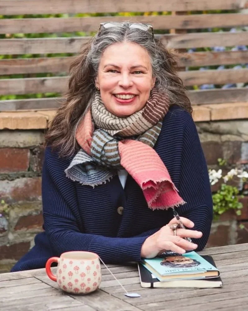 Headshot of Jane Willis smiling at the camera. Jane is sitting outside at a wooden table. A journal and mug of tea sits in front of her.