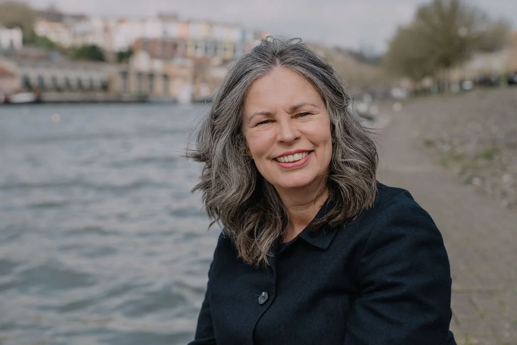 Jane Willis smiling at the camera, sitting in front of water and wearing a plain black top. Bristol harbourside is visible in the background