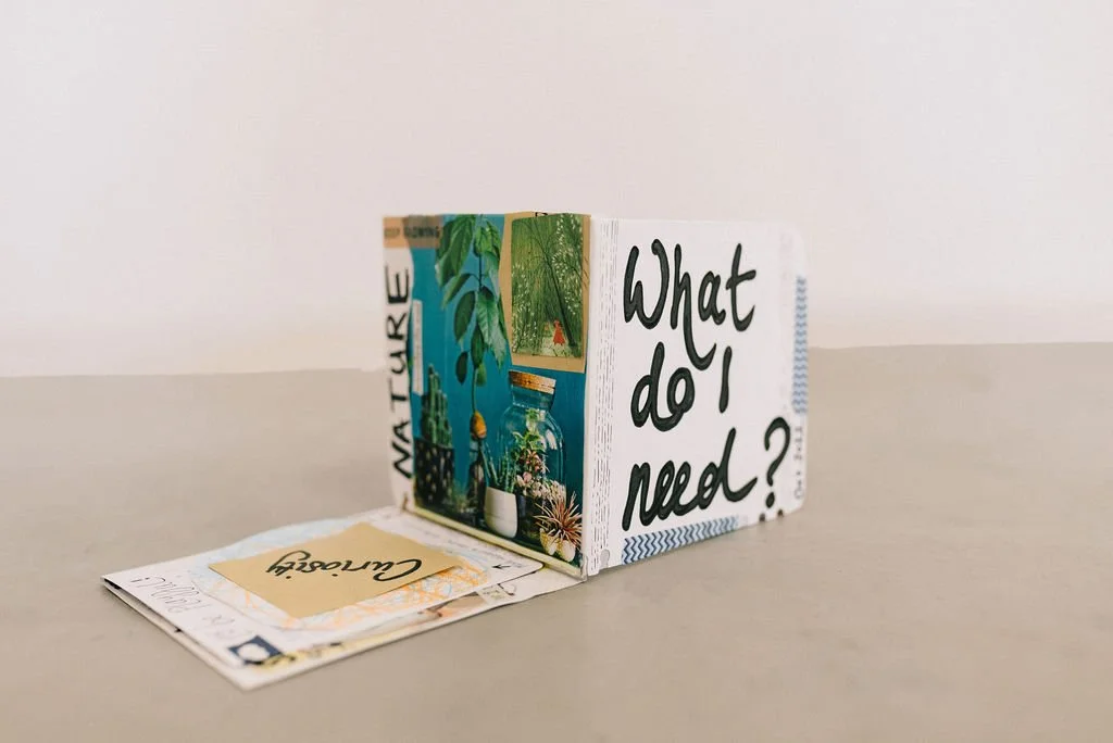 A small square handmade book with scrapbook-style features stands on a desk. The feature page on the right has the letters "what do I need" written across the page.