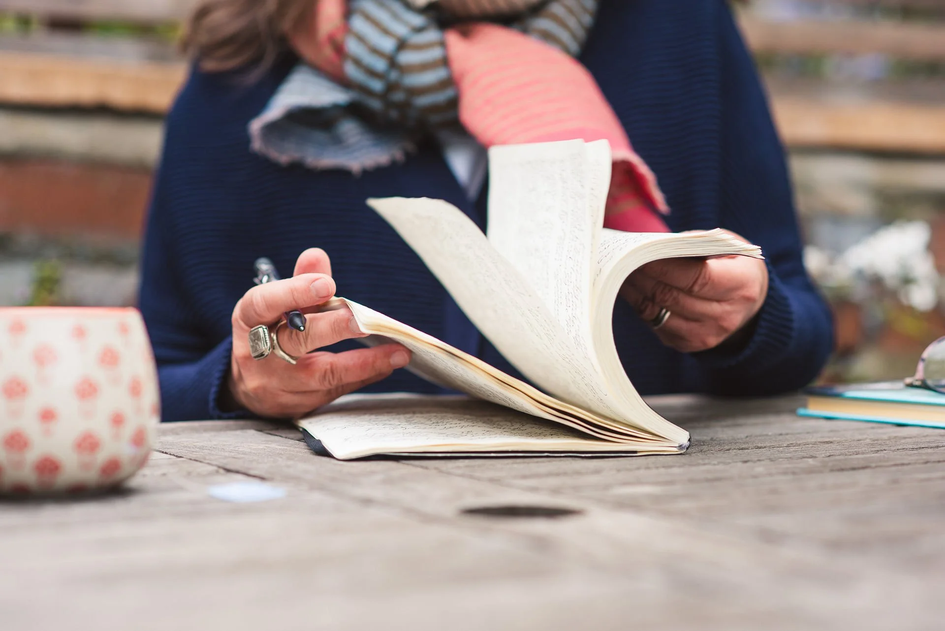 A woman flips through a journal full of writing, on a wooden table outside. A pen is in her hand, and a mug to the side.