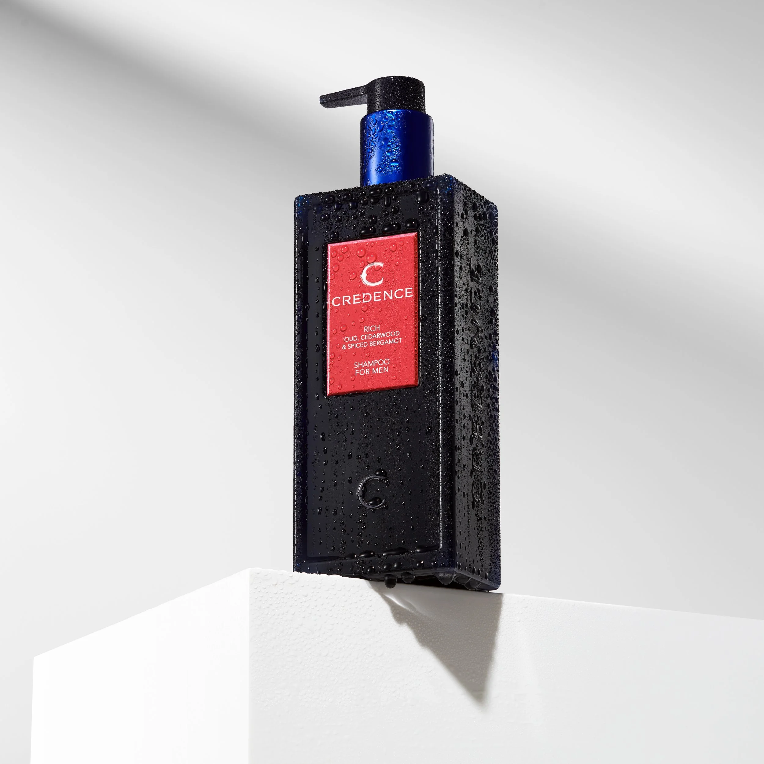 A black bottle of men's shampoo with a blue pump dispenser, water droplets on its surface, placed on a white surface against a plain background.