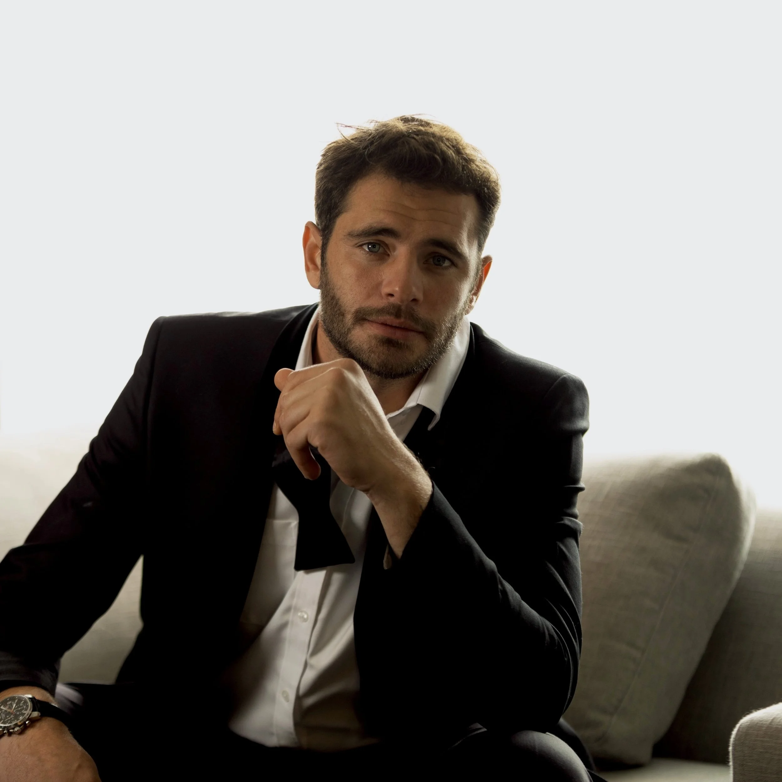 A man dressed in a black suit with a white shirt, sitting on a sofa, resting his chin on his hand, with a neutral expression.