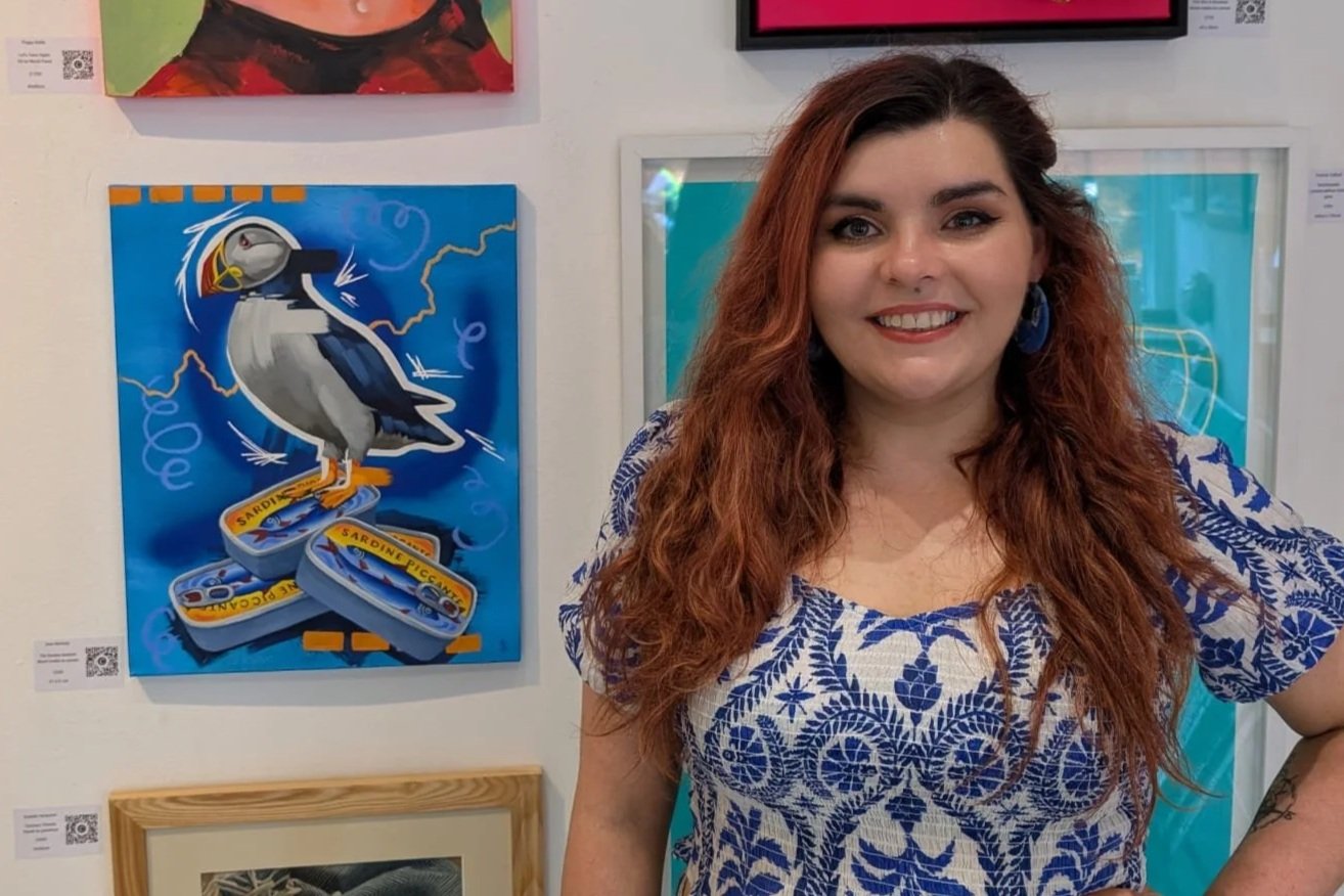 Emerging London artist Sam B. Kennedy standing beside her colourful mixed media puffin painting The Sardine Sentinel at the Everyday Magic exhibition in Archway, London.