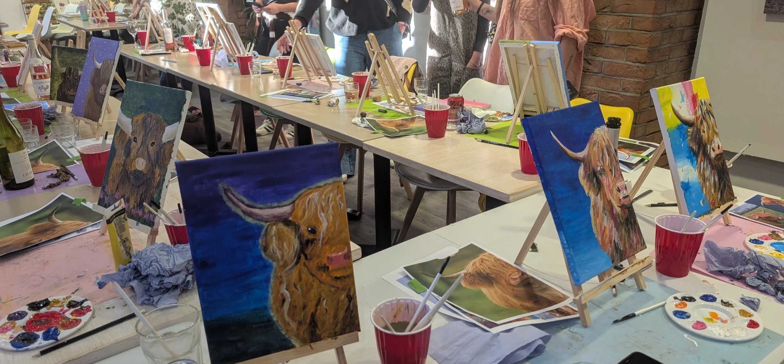 Colourful Highland cow acrylic paintings displayed on tabletop easels after a Paint & Sip art workshop in Guildford, showcasing the unique artwork created by each participant.
