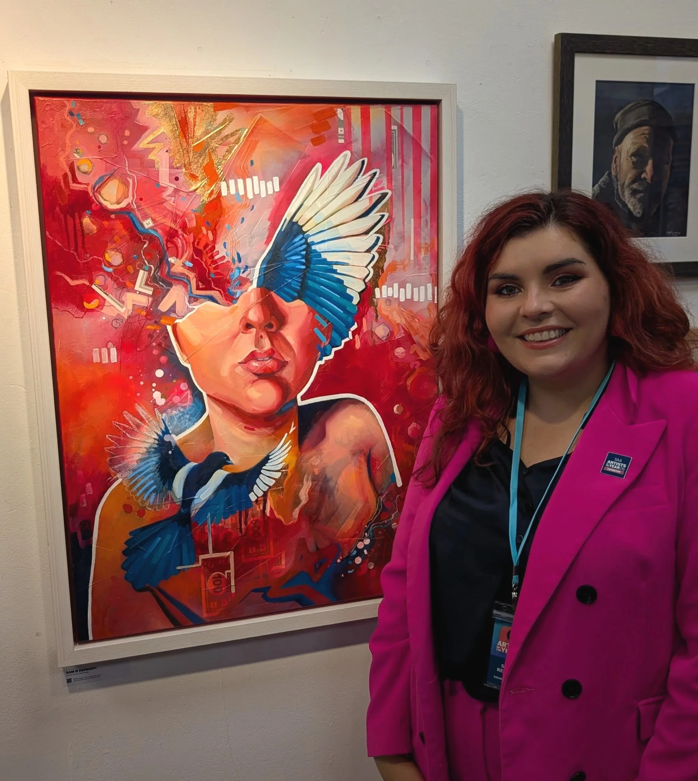 Surrey contemporary portrait artist Sam B. Kennedy standing beside Fortune Flames, her vibrant red mixed-media self-portrait in a white wooden frame, selected for the SAA Artist of the Year 2026 finalists exhibition at Cass Art Kingston.