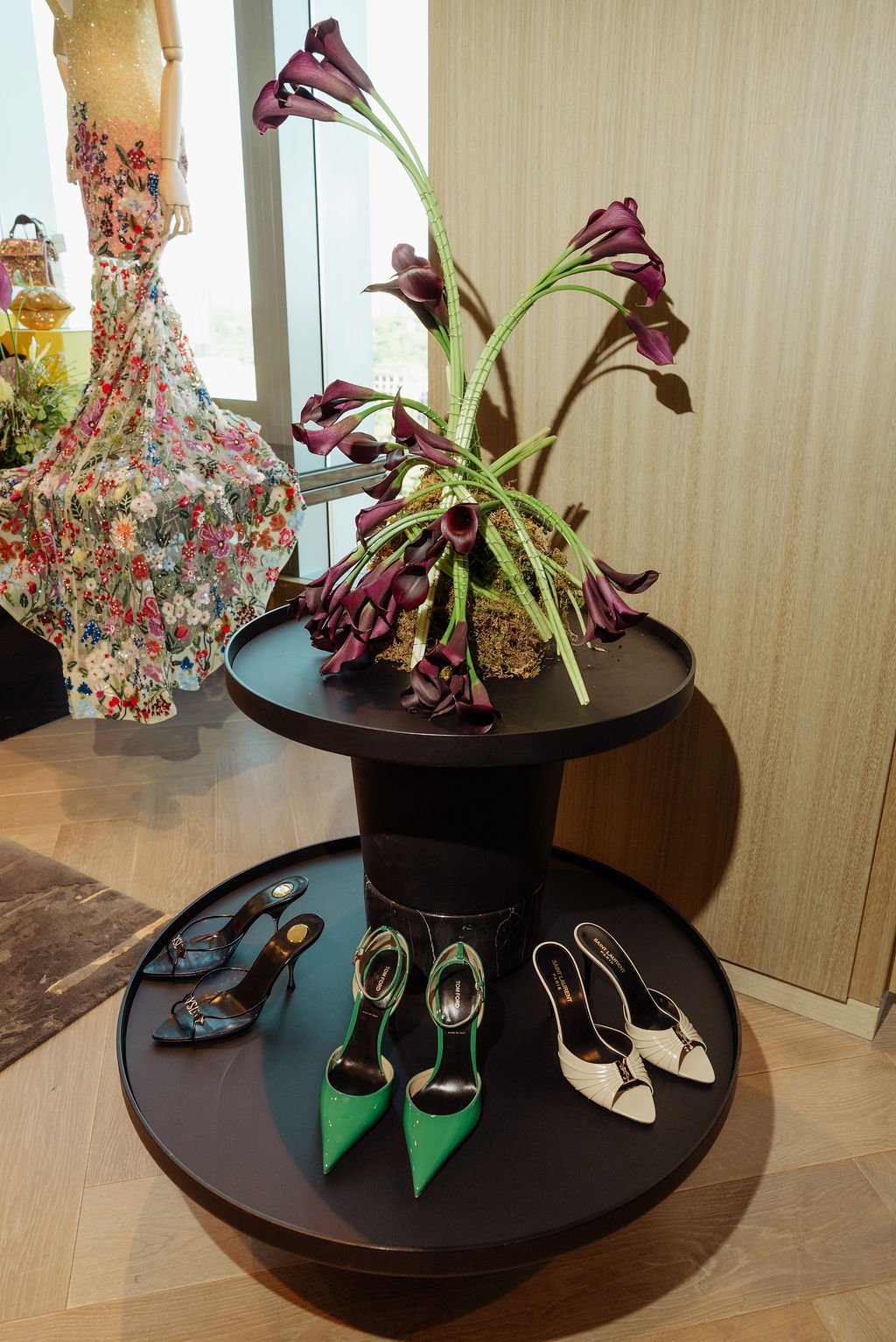 A display of high-heeled shoes on a circular black table, with a large sculptural flower arrangement, set near a window and a wooden wall.