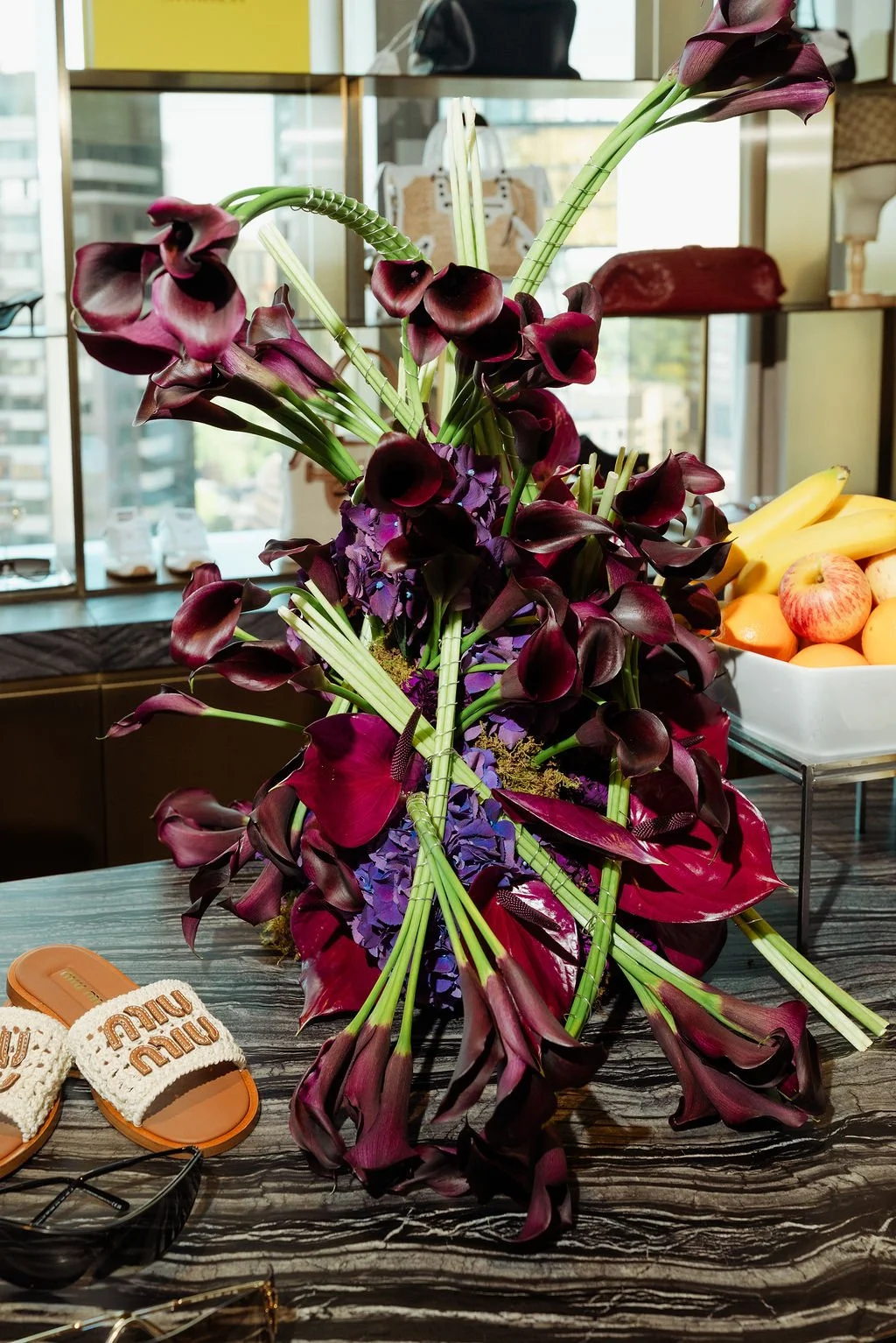 A large bouquet of dark purple calla lilies and purple flowers on a table with a fruit bowl containing bananas, apples, and oranges in the background.
