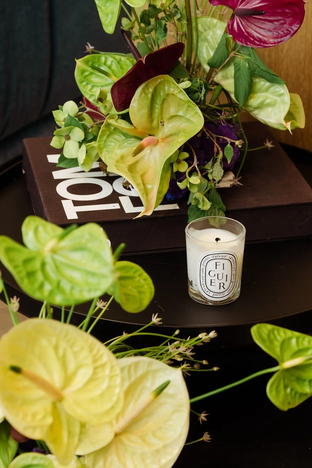 A floral arrangement of various green, yellow, and purple leaves and flowers on a black round table with a black box and a candle labeled 'FIGUE' nearby.