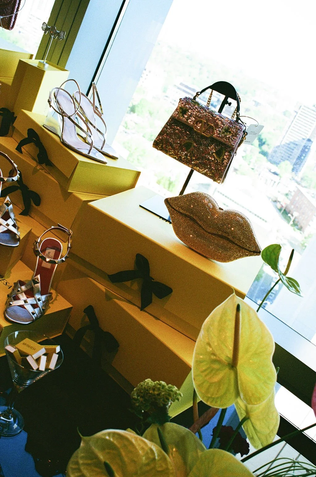 Display of luxury shoes and handbags in a retail store with a city view through the window.