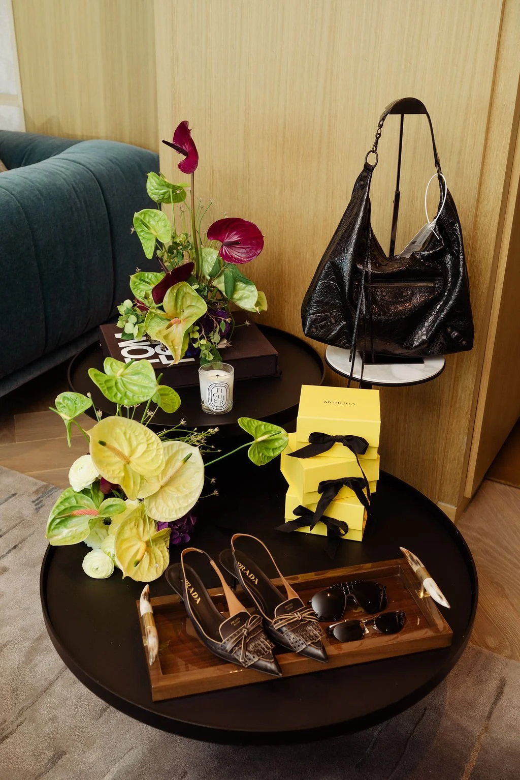 A round black table with designer shoes, sunglasses, and decorative items, including a yellow box with black ribbons, placed on a wooden tray. A black leather bag is hanging on a stand beside the table. There is a floral arrangement with green, yello