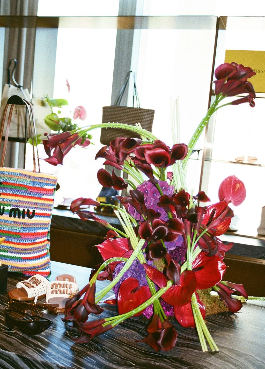 A floral arrangement with red and purple flowers on a dark wooden table in a bright room with large windows.