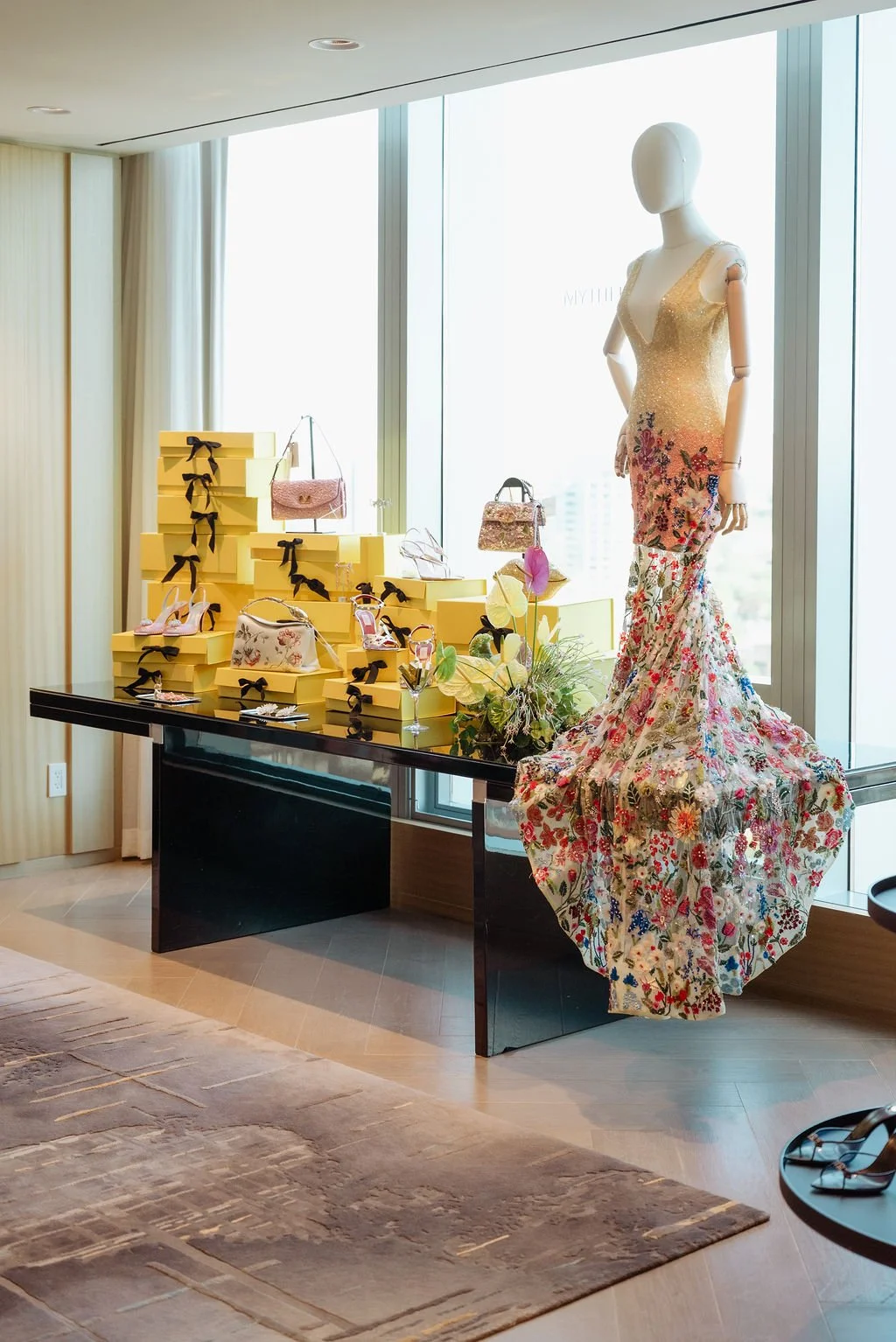 Display of high-end fashion accessories and clothing in a boutique, featuring a mannequin dressed in a floral gown, yellow boxes with black ribbons, handbags, shoes, and decorative flowers near a large window.