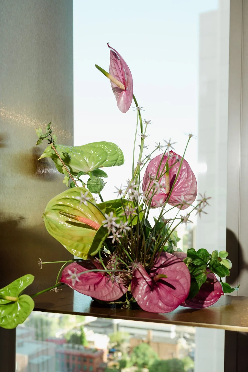 Arrangement of pink and green tropical flowers and leaves in a vase inside near a bright window.
