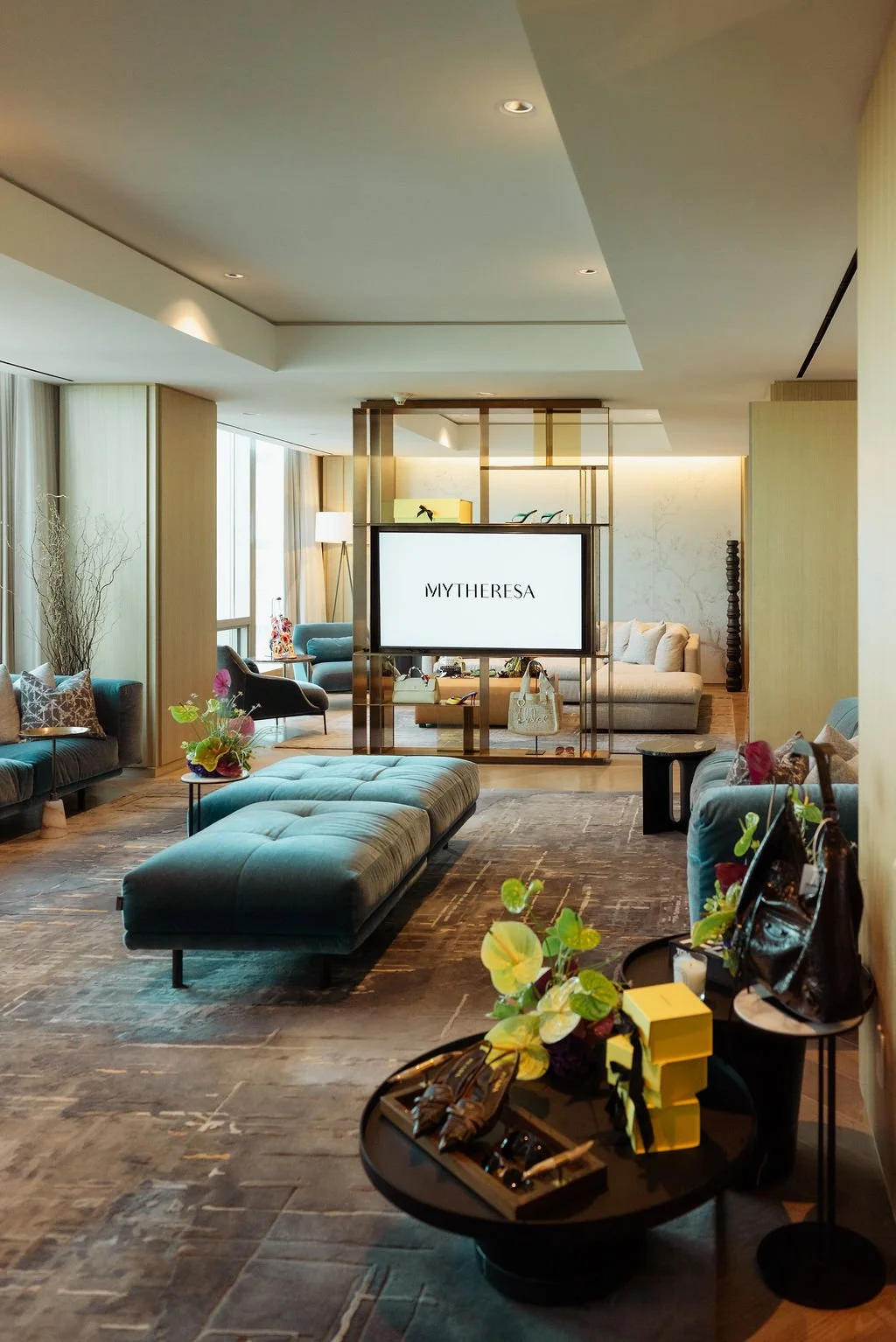 Luxurious hotel lobby with modern decor, seating area with teal and gray sofas, a large ottoman, plants, a flat-screen TV displaying "MYTHERESA," and various accessories and handbags on side tables.