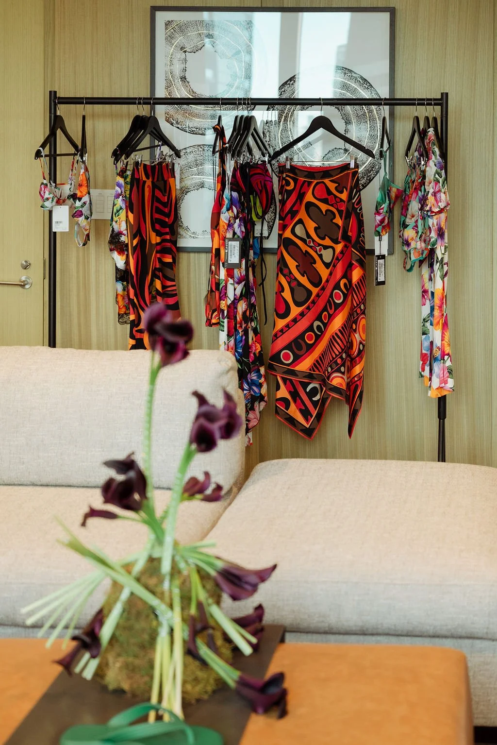 A display rack with colorful floral and patterned dresses and cover-ups behind a beige sofa and a wooden table with a purple flower arrangement in the foreground. A framed art piece is on the wall behind the rack.