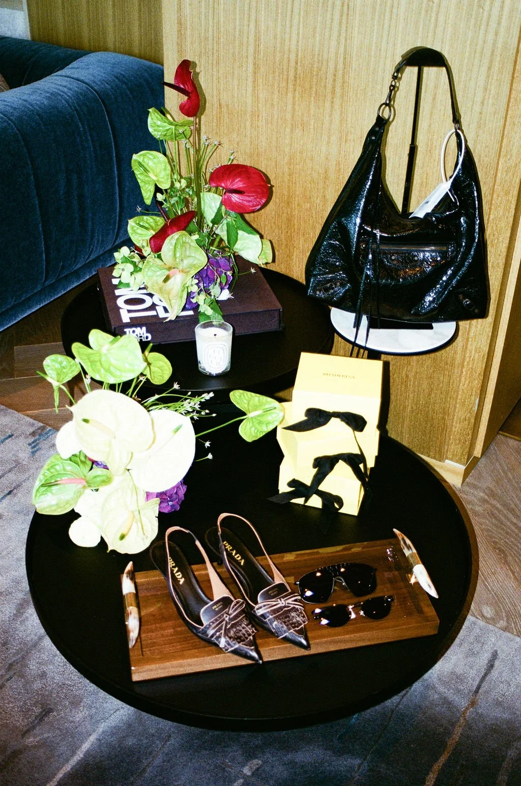 A round black table displays a pair of Prada high-heeled shoes with snakeskin pattern, black sunglasses, and an orange-tinted compact mirror. The table also has a white candle and decorative handbags, including a large black patent leather bag hangin