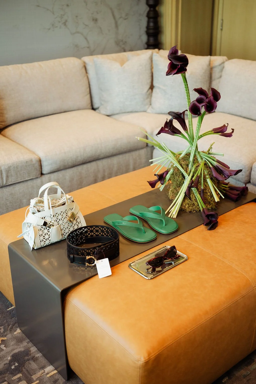 A coffee table with a large purple flower arrangement, a white handbag, green flip-flops, sunglasses, a black decorative belt, and a silver tray with sunglasses, in front of a beige sofa.