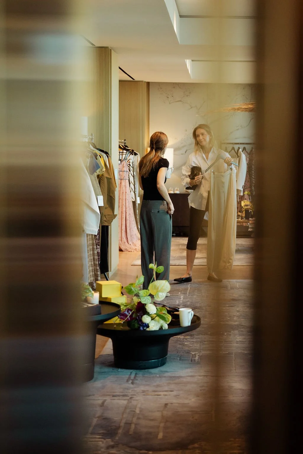 Two women are shopping for clothes in a boutique, with one showing a light-colored dress to the other. The scene is viewed through a partially blurred foreground with a black table, flowers, and cups.