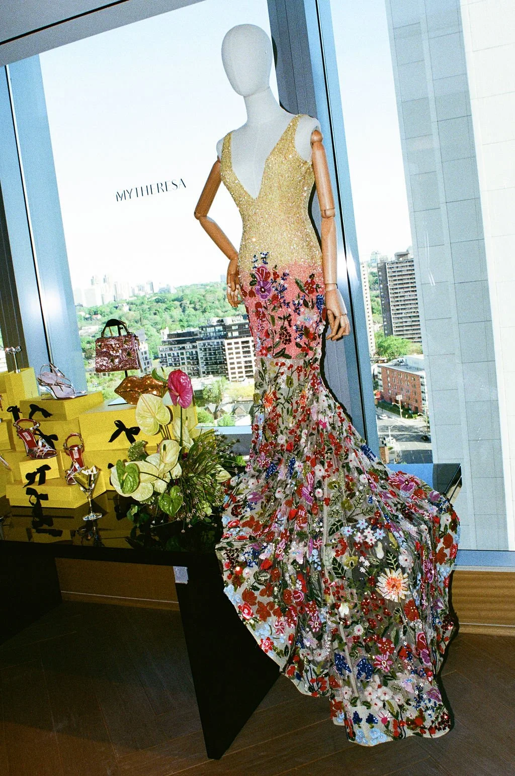 Display of designer dresses and accessories in a store window with a cityscape view in the background.
