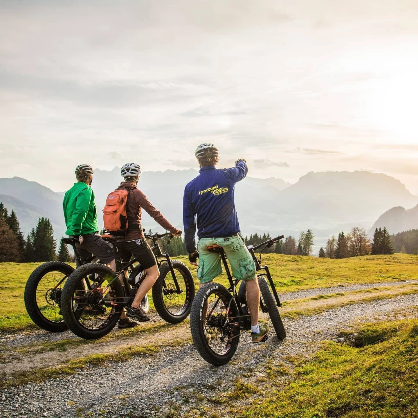 Three mountain bikers with helmets and backpacks on a trail in a lush green field, mountains in the background, during sunset.