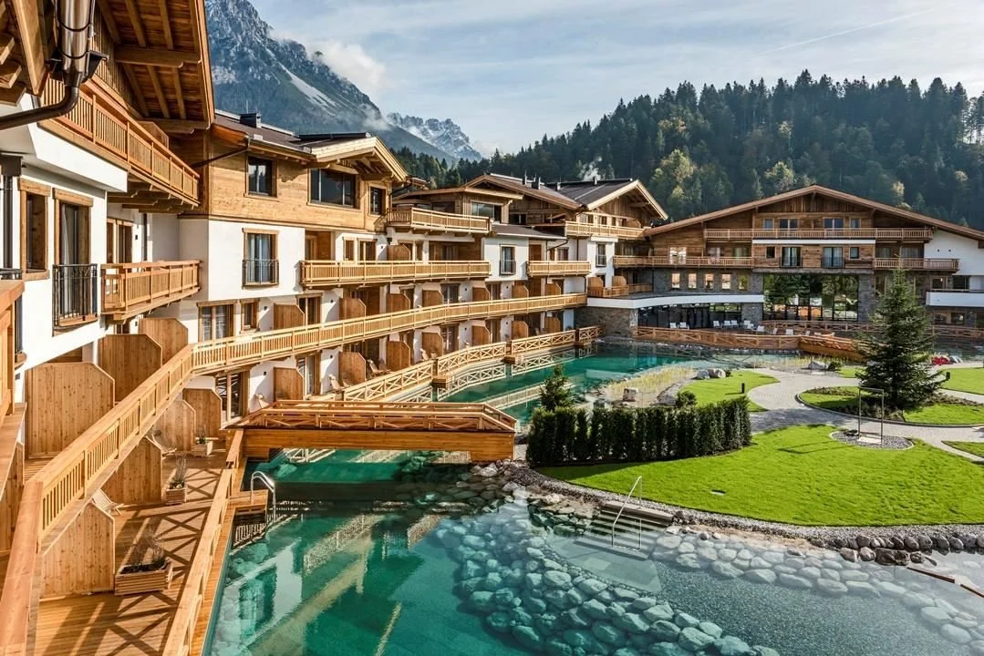 A luxurious mountain resort with wooden chalets, a swimming pool, and landscaped grassy areas surrounded by trees and mountains.