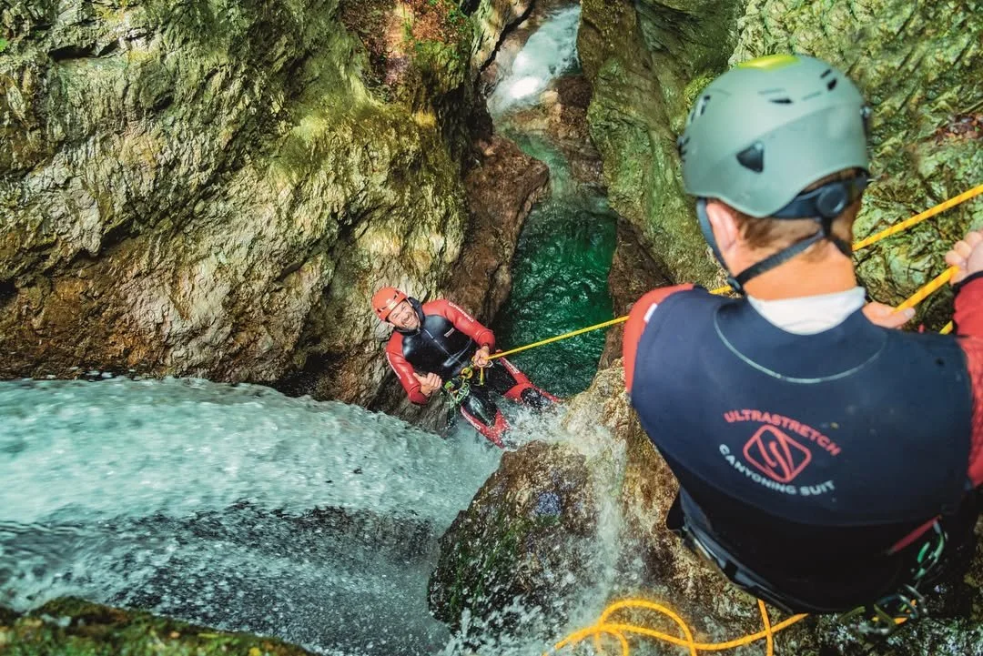 Two people canyoning in a narrow gorge with flowing water; one person is descending into the water while the other is controlling the descent from above. Both are wearing helmets and wetsuits.