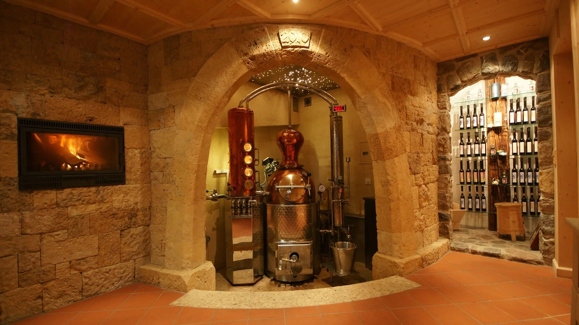 Image of a distillation apparatus in a room with stone and brick walls, a fireplace on the left, and a wine bottle display on the right.