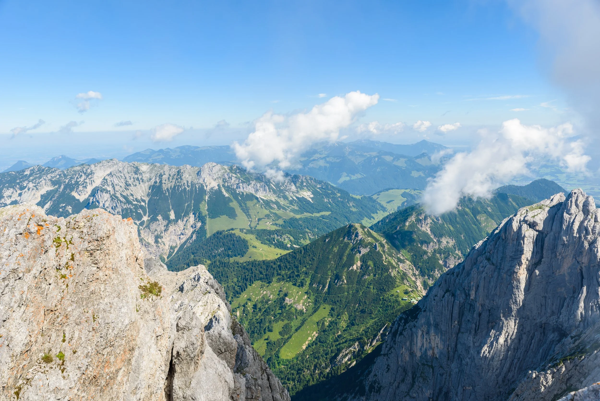 A panoramic view of a mountain range with rocky peaks, green valleys, and scattered clouds in the sky.
