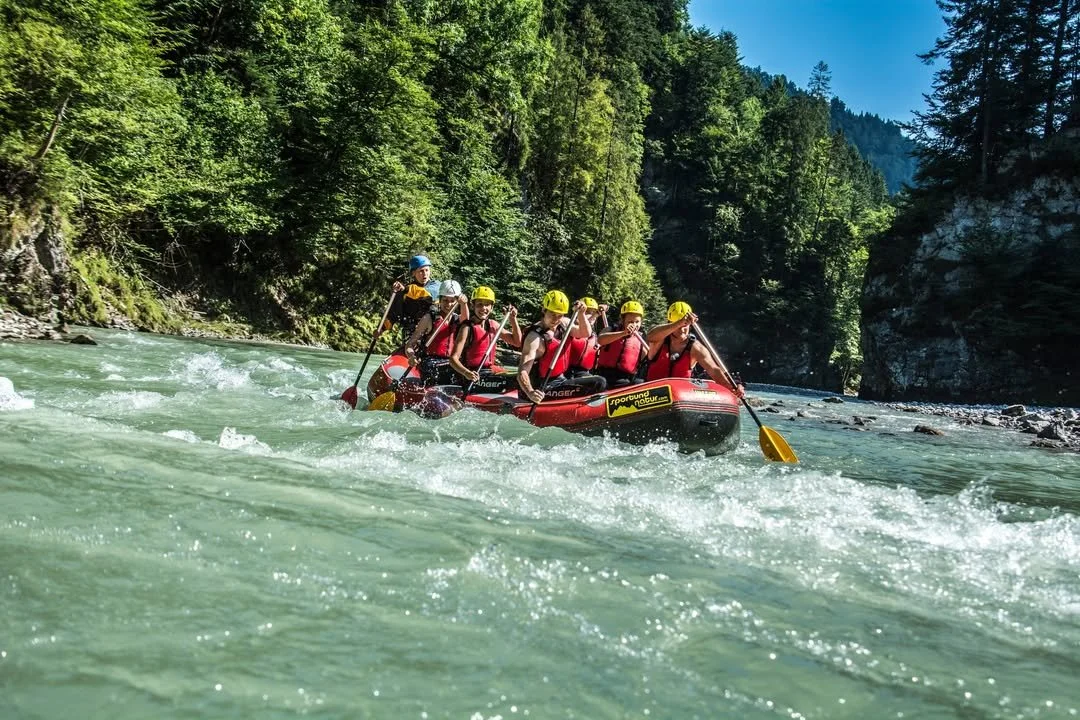Group of people white water rafting on a river with lush green trees and mountains in the background.