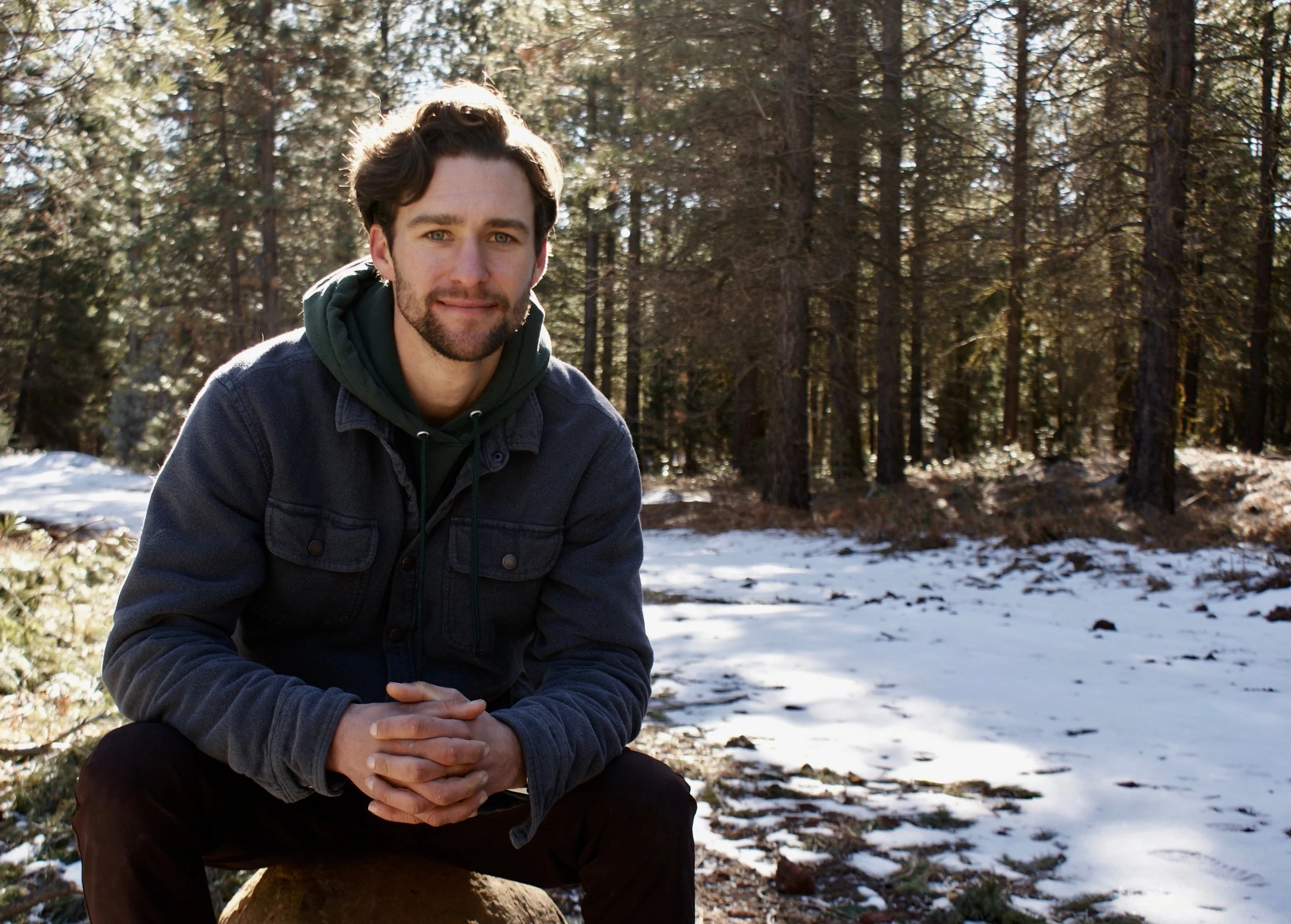 A young man with dark hair and a beard, wearing a dark jacket and hoodie, is sitting outdoors on a snowy ground with a forest of tall pine trees in the background.