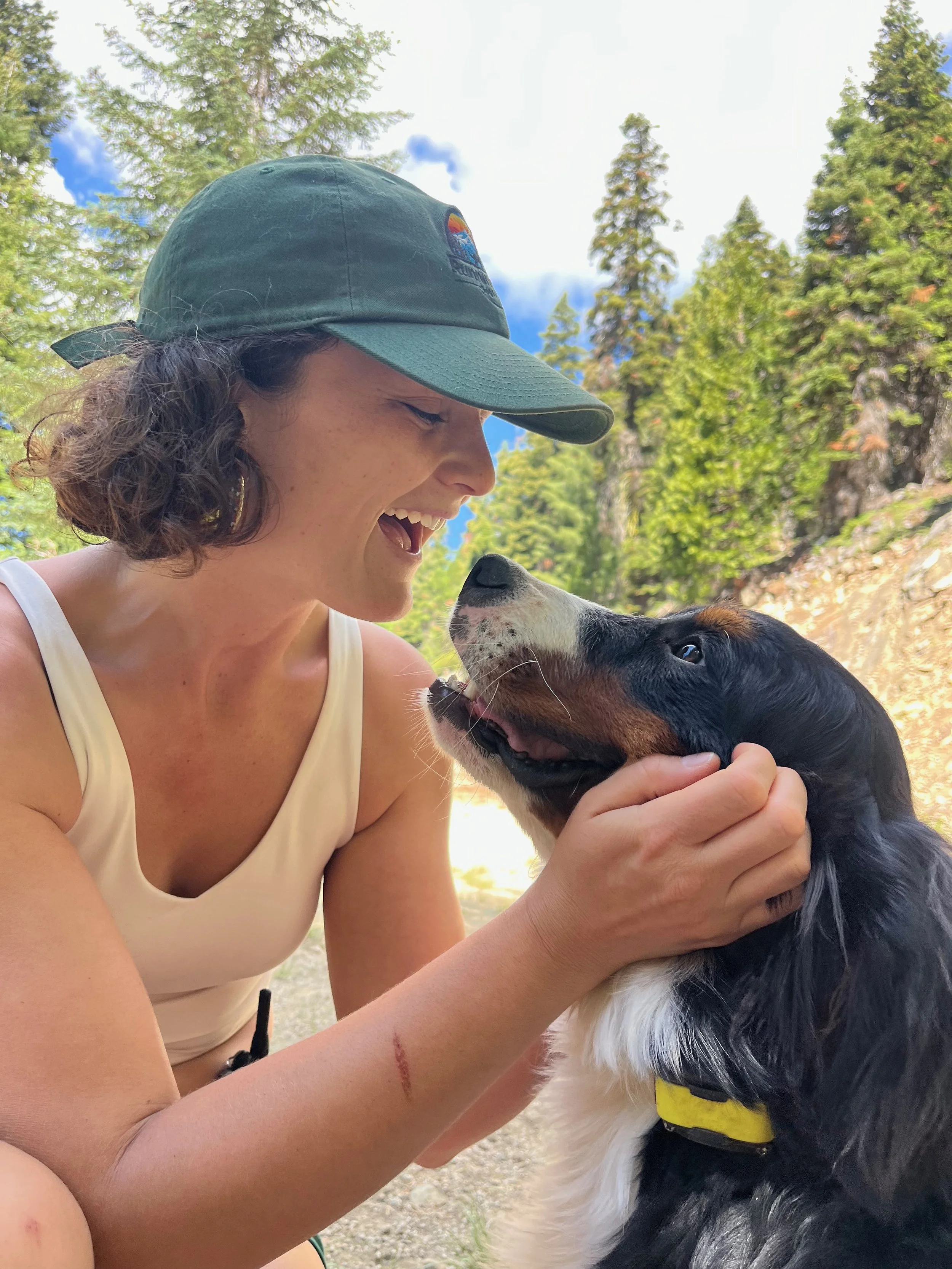 A woman with curly hair wearing a green cap smiling and leaning towards a black and white dog with a yellow collar outdoors in a forested area.