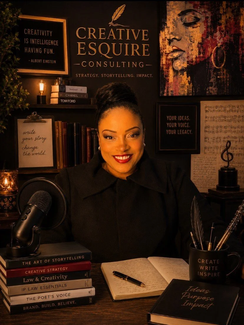 A woman with dark hair tied up, smiling in a black outfit, sitting at a desk with books, a microphone, and writing supplies, in a decorated office space with art, motivational quotes, and awards on the wall.