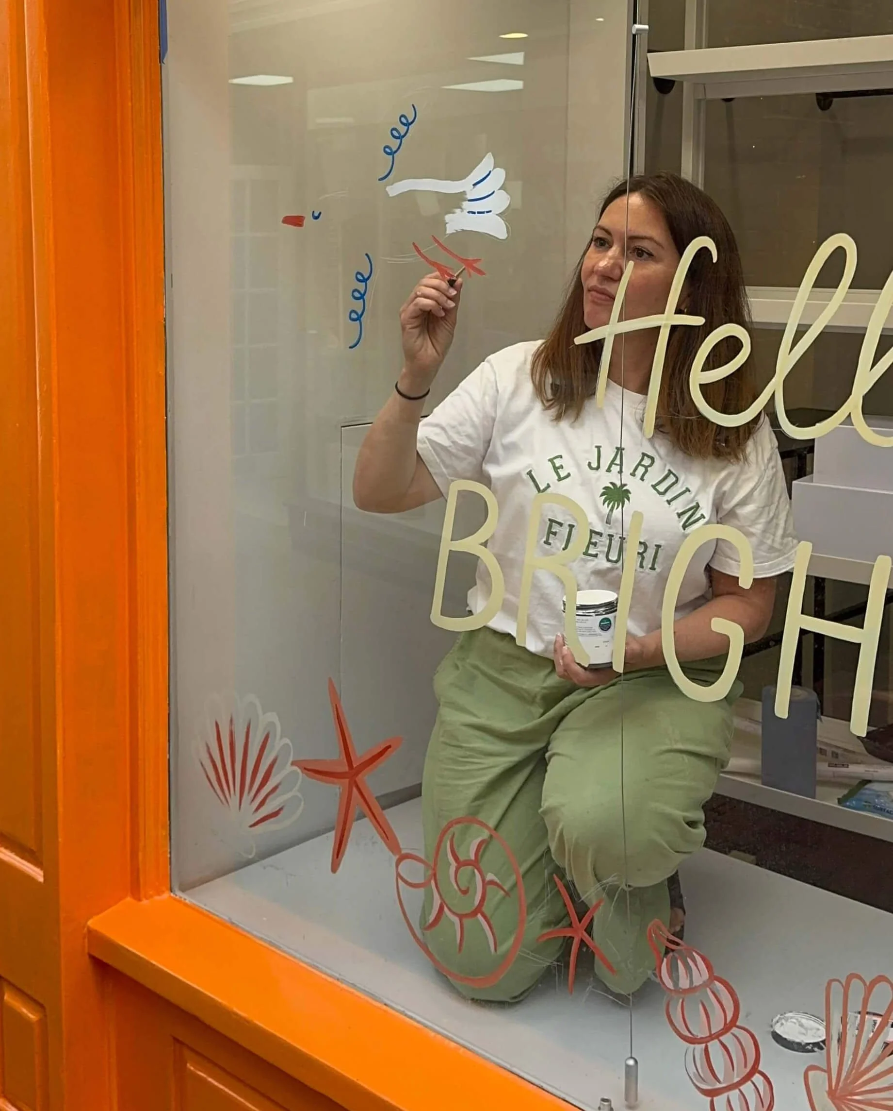 Danielle kneeling in a store window as she paints some window art