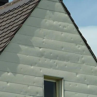 Close-up of a house's exterior wall with damage and holes in the siding, and a small window near the bottom.