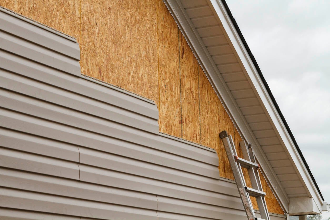 Side of a house under construction, with plywood sheathing and siding, and a ladder leaning against the wall.
