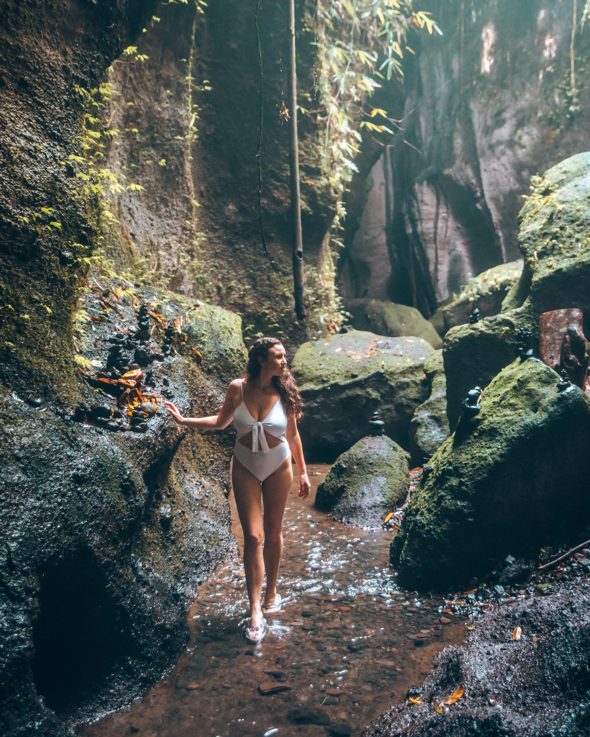 A woman in a white swimsuit walking through a small stream in a lush, rocky canyon.