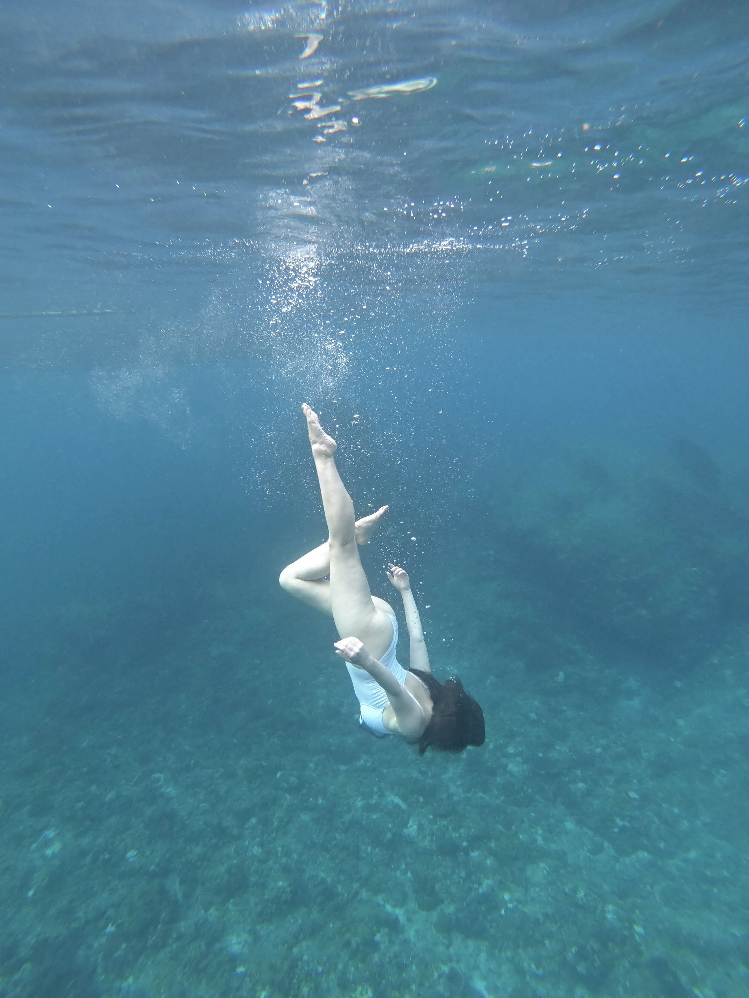 A woman swimming underwater in a white swimsuit, with bubbles and sunlight visible on the water's surface.
