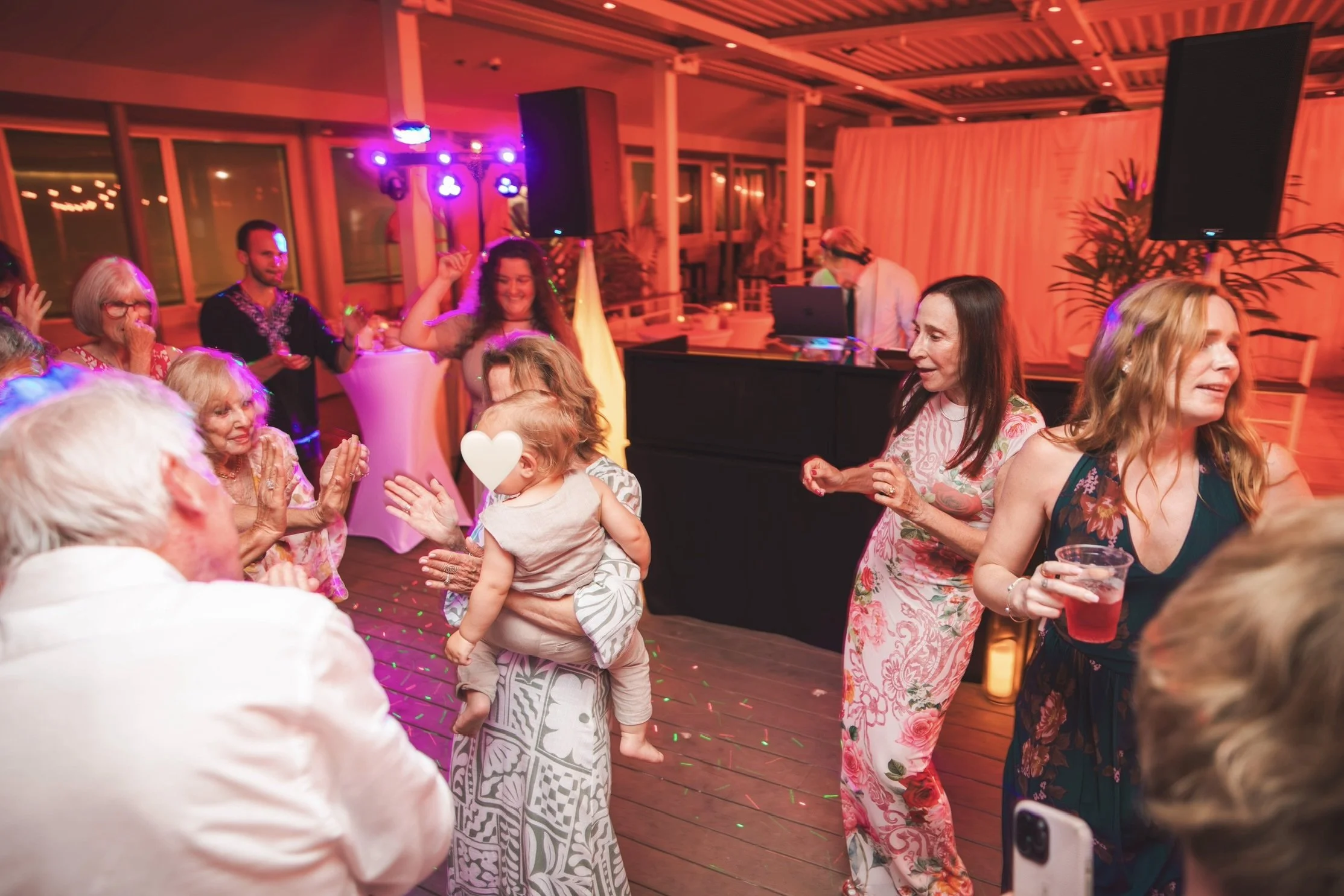People dancing and enjoying themselves at a lively party or celebration indoors, with colorful lighting and a DJ in the background.